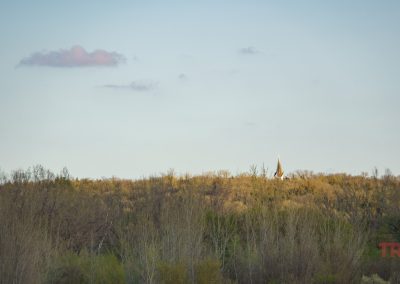 The distant church steeple of Rock Dell Lutheran Church near Belview, MN, rises above a forested landscape under a clear sky with a few clouds.