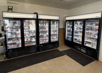 Two commercial refrigerators filled with various packaged meats sit in the corner of South Forty Meat Market, Lucan, MN. Cardboard boxes are stacked nearby at South Forty Meat Market, and certificates decorate the wall above the tile floors.