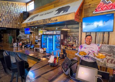 A smiling server holds plates of food at The Spotted Bear Bar and Grill in Morton, MN—a rustic spot with wooden decor, a large TV, bear-themed sign, neon lights, a well-stocked bar, and patrons relaxing in the background.