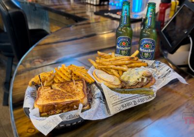 At The Spotted Bear Bar and Grill in Morton, MN, two plates await: a grilled sandwich with waffle fries and a burger topped with mushrooms and fries. Behind them sit two bottles of Heineken 0.0 beer, ready to enjoy.