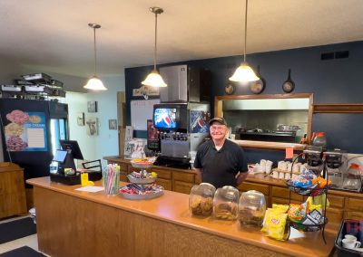 A smiling man stands behind a wooden counter at Vesta Cafe, a cozy MN diner in Vesta, MN. The counter displays cookies, chips, straws, and napkins. Behind him are coffee machines, a soda fountain, and a kitchen window. The atmosphere feels welcoming.