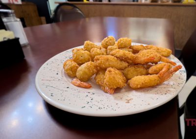 A white plate filled with golden fried shrimp and tater tots sits on a brown restaurant table at Vesta Cafe in MN, with chairs and a counter visible in the background.