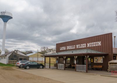 The Laura Ingalls Wilder Museum in Walnut Grove, MN, sits under a cloudy sky, with several parked cars in front and the Walnut Grove water tower visible in the background.