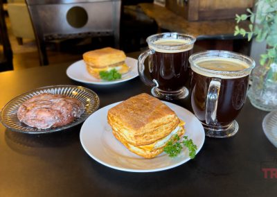 Two plates with flaky biscuits, a plate with a glazed pastry, and two mugs of dark coffee sit on a dark table in the cozy Golden Rule coffee shop in Redwood Falls, Minnesota.
