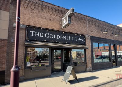 A brick storefront in Redwood Falls, Minnesota with a large black sign. The Golden Rule coffee shop features big front windows, a sidewalk sandwich board, and reflections of trees and buildings under a clear sky.