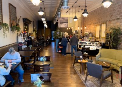 A cozy café in Redwood Falls, Minnesota, with wooden floors and tables, people dining, and a counter displaying pastries. Warm lighting at Golden Rule coffee shop, plants, and a green couch create a welcoming atmosphere amid brick walls and high ceilings.