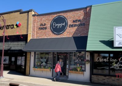 A person with a red bag walks past the brick storefront called Overjoyed with a black awning labeled Old Finds New Designs. Flower boxes decorate the window, and neighboring shops in Redwood Falls, Minnesota are visible on both sides.