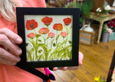 A person in a coral shirt holds a framed painting of red poppy flowers with green stems and buds. The background shows wooden floors and shelves filled with items at the Overjoyed store in Redwood Falls, Minnesota.