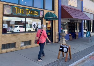 A person with gray hair walks on a sunny Redwood Falls, Minnesota sidewalk toward the Redwood Area Arts store. The storefront features large windows, a green awning, and a sidewalk sign that reads OPEN TODAY.