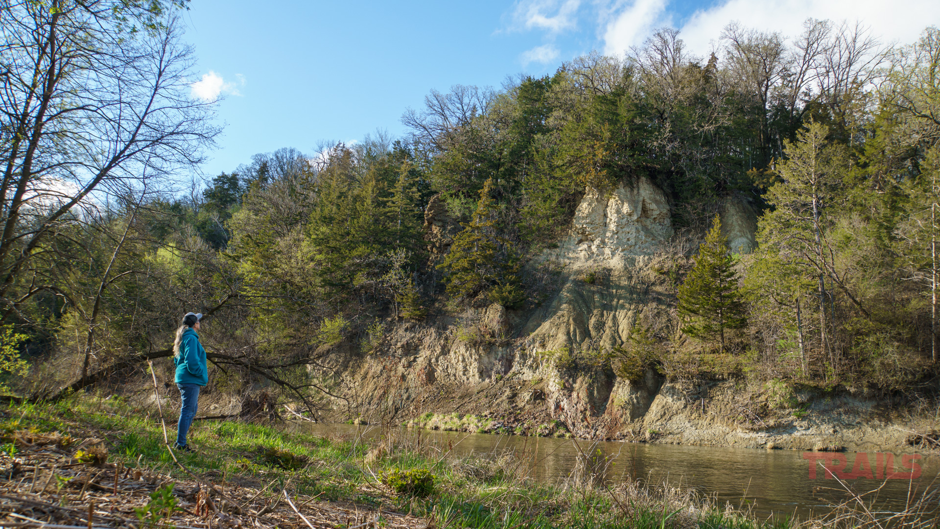 Redwood River Bluffs Cansayapi Park A person stands on grassy ground by the Redwood River in Ramsey Park/Cansayapi, Redwood Falls, MN, gazing at a steep, tree-covered hillside under a partly cloudy blue sky.