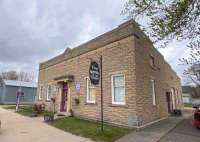 Fond Memories in Walnut Grove is housed in a rectangular brick building on a corner in Walnut Grove, MN. It has red doors, a blue historical marker, and a cloudy sky above. Cars are parked along the street, and trees are visible nearby.