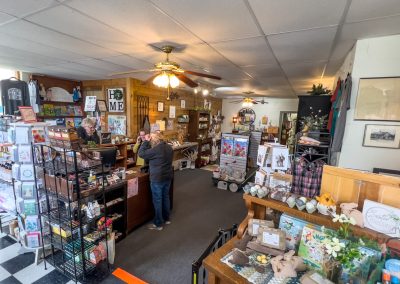 Small gift shop interior in Walnut Grove with shelves and displays of various items, two people at the counter, checkered floor, and decorations throughout the store, including books, signs, home decor, and Fond Memories stamp shop exclusives.