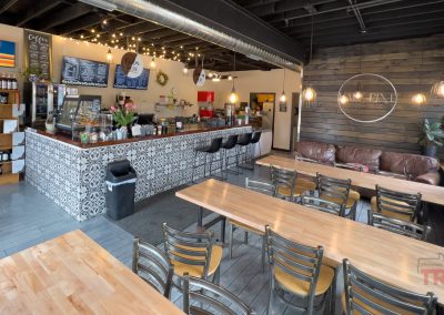 Half Pint Coffee Co. is a cozy coffee shop with wooden tables, metal chairs, a patterned counter, a menu on the wall, shelves of coffee products, string lights, and a seating area featuring a brown leather couch and plants.