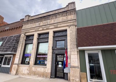 A historic stone building with BANK carved above the entrance stands between two modern storefronts. An Open flag hangs by the door under a cloudy sky.