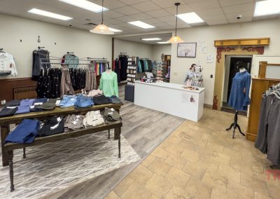 Plum Creek clothing boutique interior with racks of shirts, dresses, and jackets, a table displaying folded jeans and tops, and a white counter with a cash register. The space has light walls, hanging lights, and tiled floors.