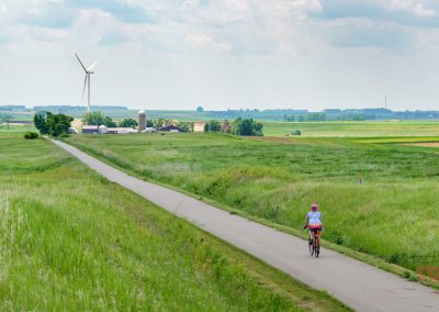 A person rides a bicycle on the paved Casey Jones Trail through green fields, passing a wind turbine and farm buildings in the distance under a partly cloudy sky.