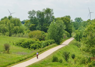 A person rides a bike on a paved path through green fields and trees along the Casey Jones Trail, with two wind turbines visible in the background under a partly cloudy sky.