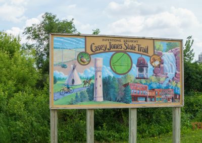 A colorful sign for the Pipestone Segment of the Casey Jones State Trail, featuring paintings of a cyclist, a windmill, buildings, a waterfall, a train, and various local landmarks, set near green bushes and a partly cloudy sky.