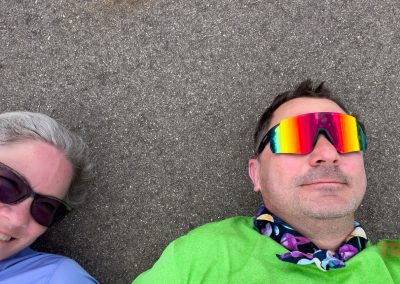 A smiling woman with gray hair and sunglasses lies next to a man in rainbow sunglasses and a green shirt, both on a gray pavement. Only their upper bodies and faces are visible.
