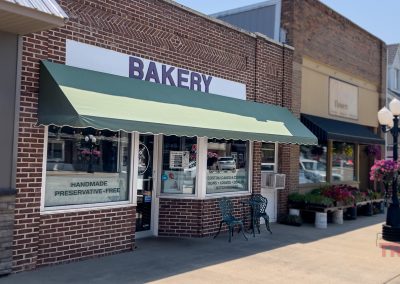 A brick bakery storefront with a green awning, outdoor seating, and a sign reading HANDMADE PRESERVATIVE-FREE. Colorful flowers hang by nearby shops along the sunny Edgerton sidewalk at Edgerton Bakery.