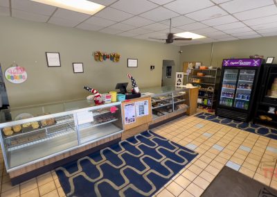 The interior of Edgerton Bakery in Edgerton MN features empty glass display cases, a counter with a register, drink coolers, shelves of snacks and drinks, tiled floors, and walls adorned with decorations and menu signs.
