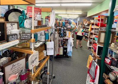 A brightly lit Katies Closet store aisle in Edgerton MN is filled with shelves of gifts, mugs, signs, kitchenware, and various novelty items. A person in shorts browses the displays further down the aisle.