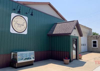 A green metal building with a small entrance canopy displays a round sign reading Spronk Artisan Bakery. A glass display case with plants and flowers sits outside on the sidewalk in Edgerton MN.