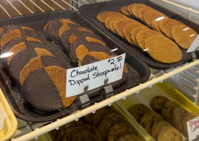 Trays of cookies on display in a bakery case at Spronk Bakery in Edgerton MN, featuring chocolate-dipped stroopwaffels in the foreground with a handwritten label and price, and other cookies arranged nearby on shelves.