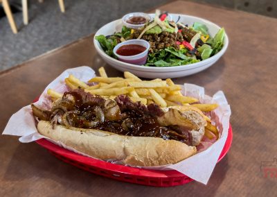 A basket with a sandwich topped with grilled onions and sauce, served with fries. In the background, a salad with greens, vegetables, and dressing sits in a white bowl on a brown table at Tally Ho Koffie in Edgerton MN.