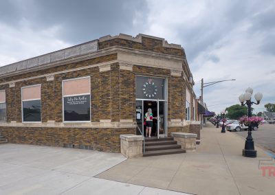 A brick corner building in Edgerton MN, once a “BANK,” now hosts Tally Ho Koffie cafe. People enter as vintage street lamps with flower baskets line the sidewalk on a cloudy day.