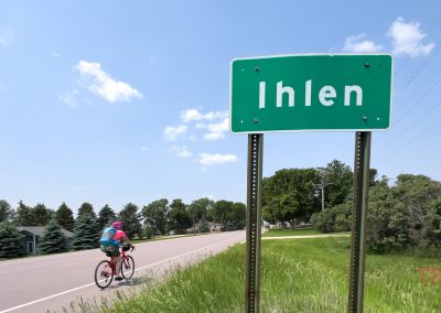 A cyclist rides on a rural road past a green sign that reads Ihlen under a blue sky, with houses, trees, and grass in the background.