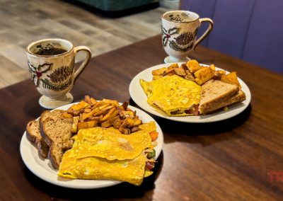 Two plates of breakfast omelets with toast and home fries, each paired with a patterned mug of coffee, are set on a wooden table at Fish and Chips Restaurant in Ihlen MN. The food looks fresh and warm, ready to be enjoyed.