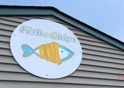 A round sign on the Fish and Chips Restaurant in Ihlen MN features a cartoon fish wrapped in fries, with the text Fish & Chips in yellow and blue letters above it.