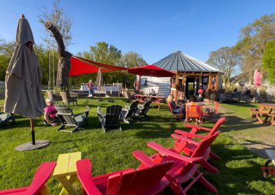 Outdoor seating area with colorful Adirondack chairs on grass, people relaxing near the 1910 Sip House in Battle Lake, MN. Features a round gazebo-like structure, red sunshades, picnic tables, and trees under a clear blue sky.