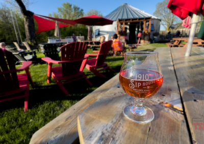 A glass of cider sits on a wooden table outdoors near red Adirondack chairs at 1910 Sip House in Battle Lake, MN, with people and a rustic building in the background on a sunny day.