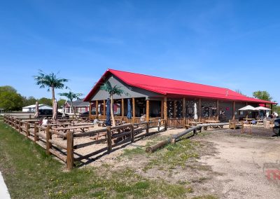 A building with a red roof and wooden outdoor seating, home to ABC Brewing, is surrounded by a wooden fence on a sunny day in Battle Lake, MN. A sidewalk and parked vehicles are nearby, with trees and blue sky in the background.