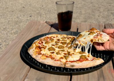 A hand lifts a cheesy slice of meatball pizza from a black tray on a wooden table outdoors in Battle Lake, MN. A glass of dark beer from ABC Brewing rests in the background.