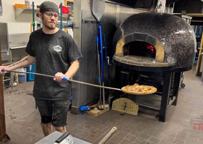A man wearing a black cap and shirt uses a long-handled peel to remove a pizza from a wood-fired oven in the ABC Brewing kitchen in Battle Lake, MN. The oven has a dark, mosaic-tiled dome and a visible fire inside.