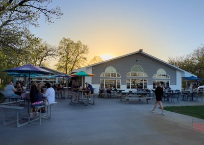 People sit and eat at outdoor tables under colorful umbrellas outside the light gray Beach Bums bar in Battle Lake, MN at sunset. Trees and a clear sky are in the background, with a few people walking nearby.