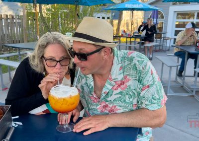 A man and woman share a large orange cocktail at Beach Bums bar in Battle Lake, MN, sitting close together on the outdoor patio. The man wears a tropical shirt and straw hat; the woman wears glasses. Others relax at picnic tables nearby.