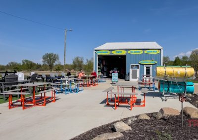 Outdoor seating area with colorful benches and tables in front of a small blue building in Battle Lake, MN, with people relaxing, a large roll of hay, barrel equipment nearby, and Big Fish Kayaks surrounded by greenery and rocks.