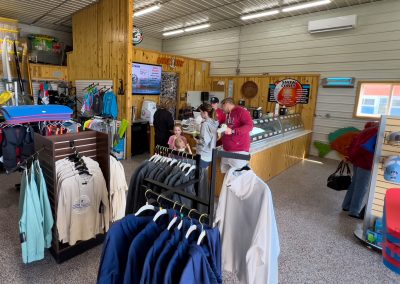 A store interior in Battle Lake, MN, with wooden walls and racks displaying clothing, hats, water sports gear, and Big Fish Kayaks. Several people gather at the counter beneath a menu board. Bright lighting fills the spacious, organized shop.