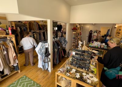 Inside MN Chelsea Pacifica Boutique in Battle Lake, racks of clothes, shelves of hats, and jewelry displays fill the cozy, well-lit space. Two women browse the merchandise as another stands near the counter.