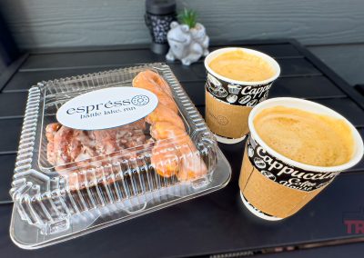 Two cups of coffee from Esprésso coffee shop in Battle Lake, MN, and a clear plastic container with pastries sit on a black outdoor table. A small decorative frog and a plant are in the background.