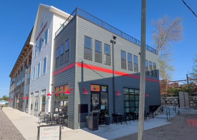A modern, two-story gray building with red accents in Battle Lake, MN features large windows and outdoor seating. The Esprésso coffee shop sits along a sidewalk on a sunny day with clear blue sky.