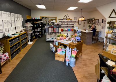 A cozy boutique in Battle Lake, MN, Funky Farmhouse features wooden floors and displays of home décor, mugs, towels, and gifts. A woman sits at a desk in the back. The walls are adorned with framed art and shelves of unique items.