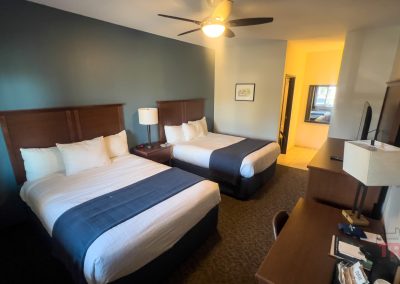 Hotel room at Battle Lake Inn motel in Battle Lake, MN, featuring two double beds with dark wood headboards, white and navy bedding, a ceiling fan, desk with chair, TV, lamp, and a small bathroom visible through an open door.