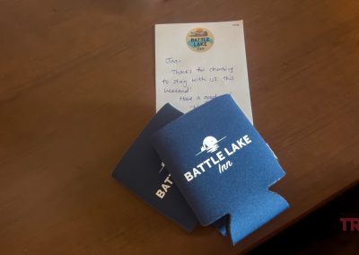 A handwritten note and two blue Battle Lake Inn motel can coolers rest on a wooden table. The note thanks a guest for staying in Battle Lake, MN.