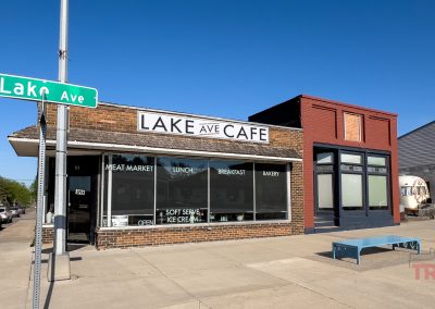 A brick building on a street corner in Battle Lake, MN, houses the Lake Avenue Cafe, with large front windows displaying menu items. A green Lake Ave street sign is on the corner, and a blue bench sits on the sidewalk.
