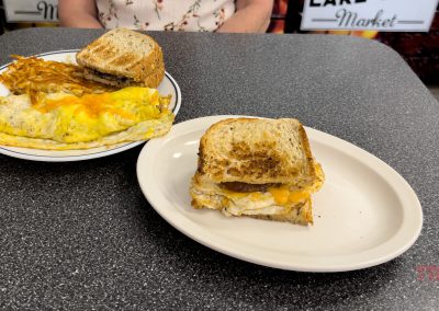 A plate with a fried egg and cheese sandwich on toasted bread is in the foreground at Lake Avenue Cafe. In the background, another plate holds an omelette with melted cheese, hash browns, and toast in Battle Lake, MN. A person is seated behind the plates.